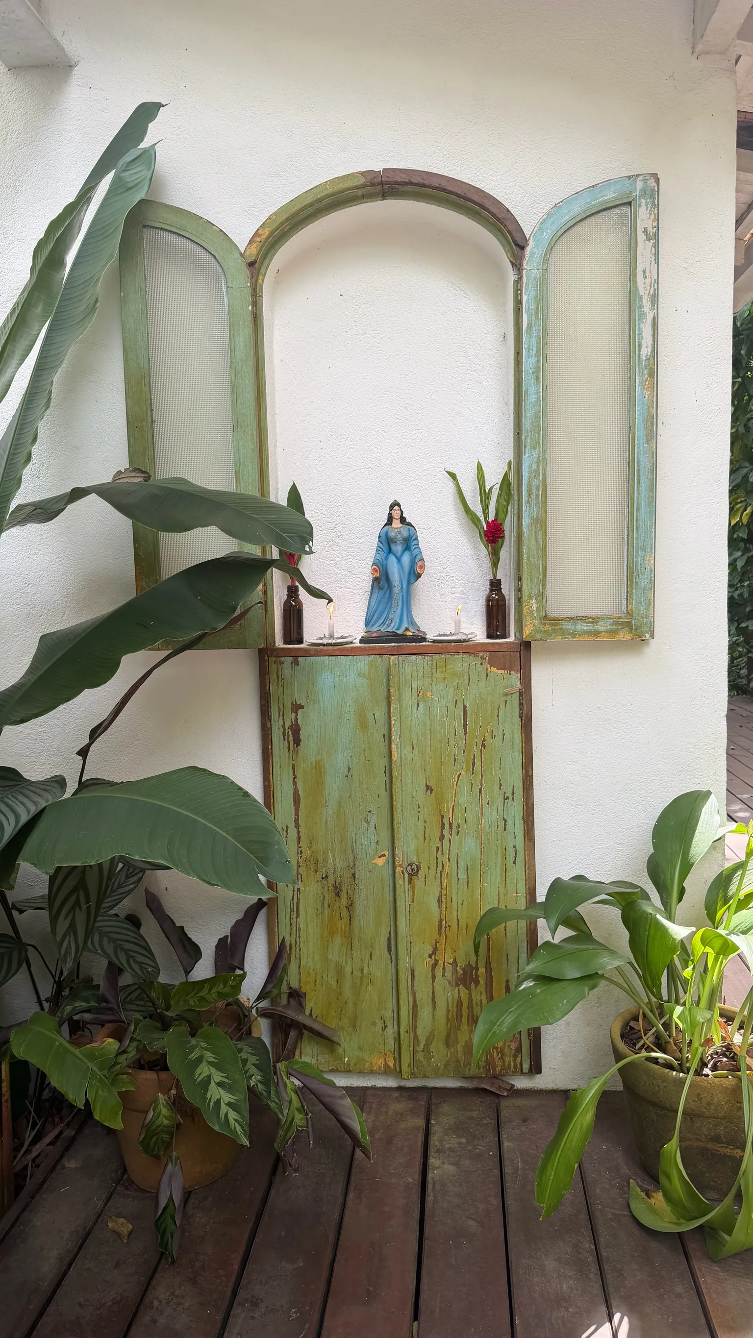 Oratório de madeira descascada encaixado na parede branca, com velas acesas e plantas em volta.